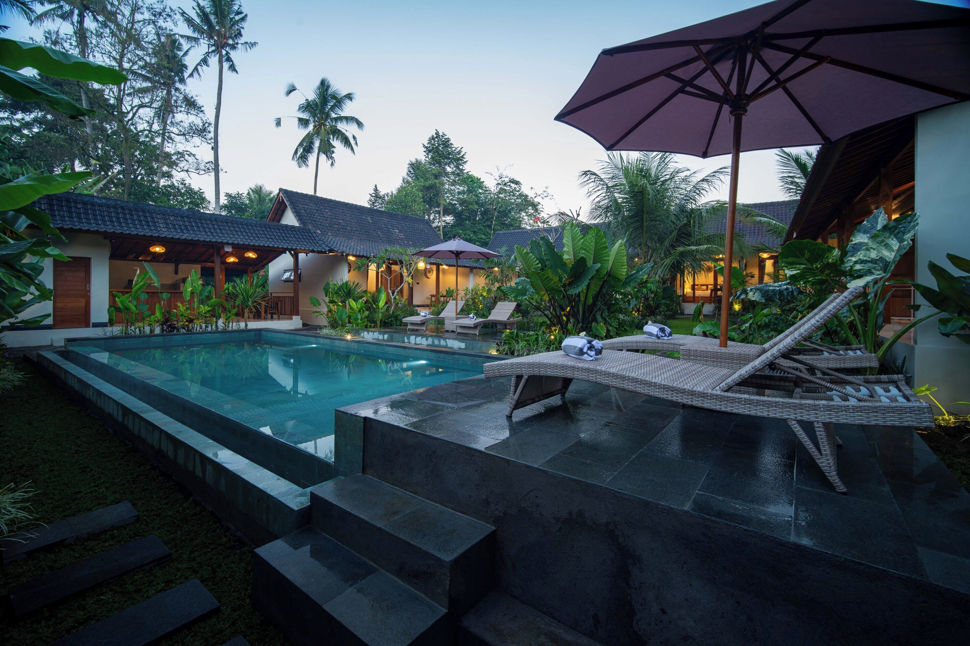 Uma área de piscina com espreguiçadeiras e guarda-sóis, rodeada por plantas tropicais e edifícios com telhados de azulejos.