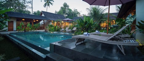 A pool area with lounge chairs and umbrellas, surrounded by tropical plants and buildings with tiled roofs.