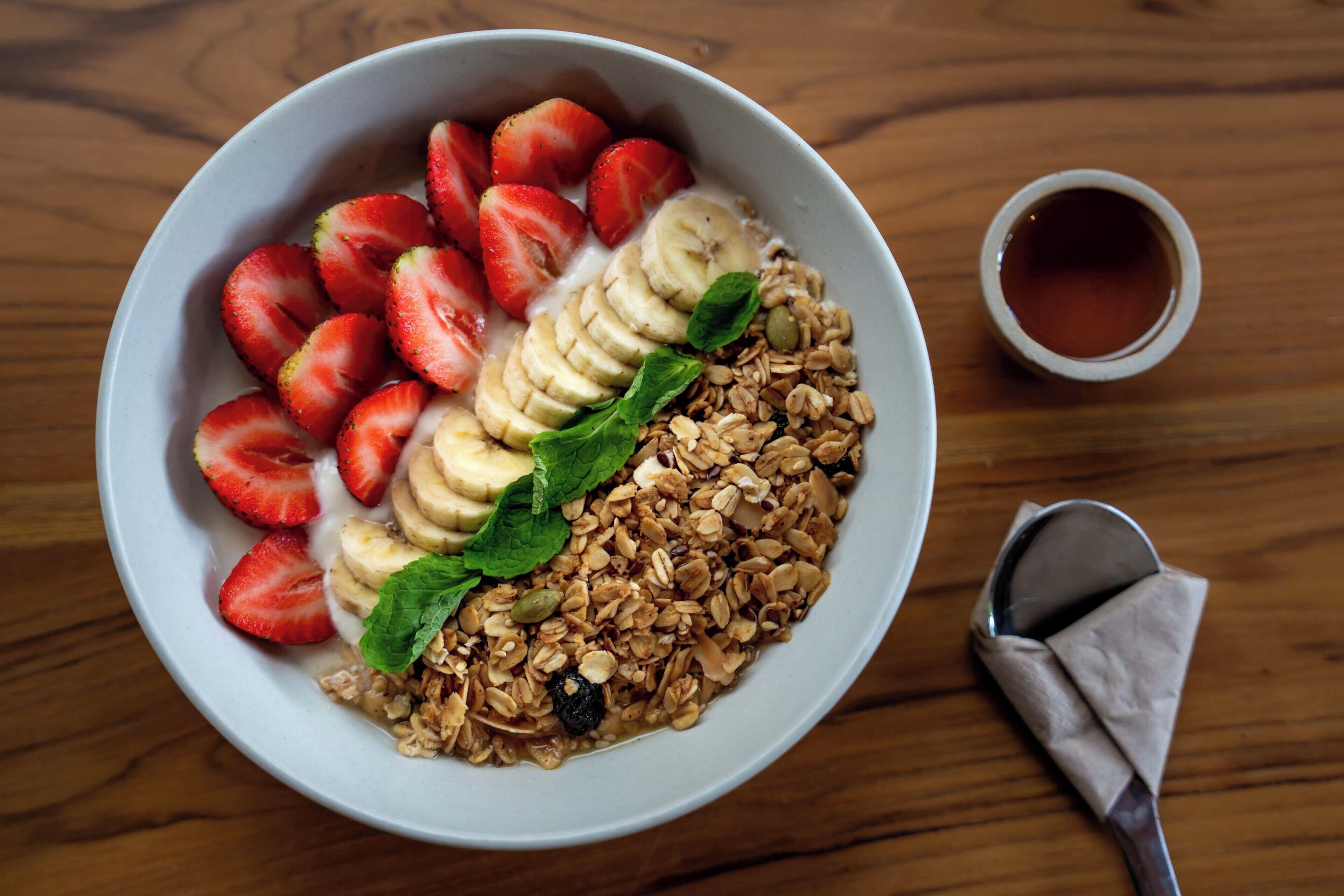 A bowl of cereal with strawberries and banana slices, a cup of tea, and a spoon on a wooden table.