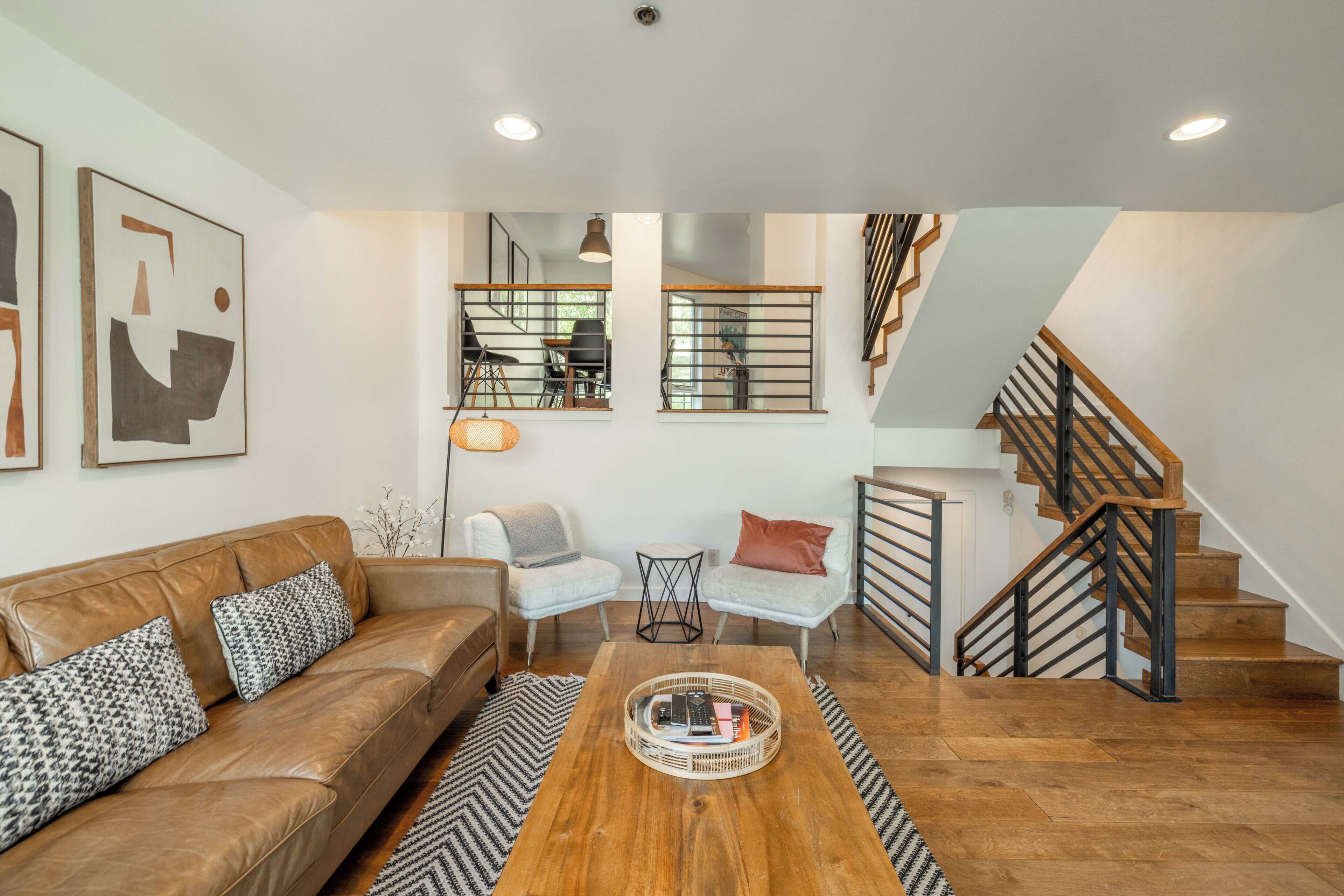 A modern living room with a brown leather sofa, two armchairs, a coffee table, and a staircase with wooden railings.