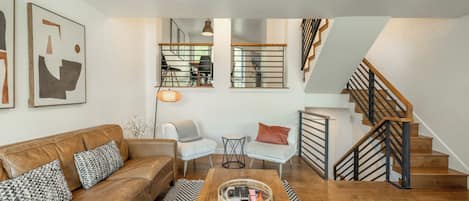 A modern living room with a brown leather sofa, two armchairs, a coffee table, and a staircase with wooden railings.