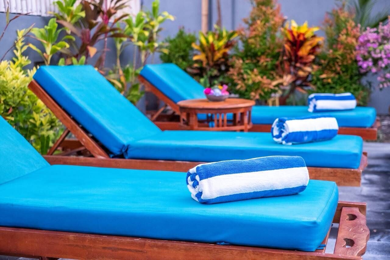 Una hilera de sillones de madera con cojines y toallas azules, dispuesta al aire libre con plantas y una pequeña mesa al fondo.