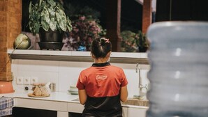 A person in a red and black uniform working at a kitchen counter with a sink and a potted plant in the background. - The Lonceng Canggu (Canggu)
