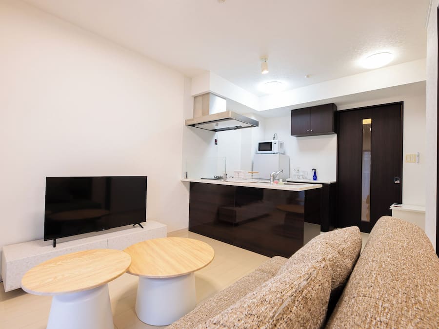 A modern living room with a flat-screen TV, two round wooden tables, a beige sofa, a kitchen area with a microwave, and a dark wooden door.