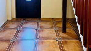 Un pasillo con una puerta oscura que incluye un vitral, una escalera de madera con barandilla roja y un suelo de baldosas con un diseño geométrico.