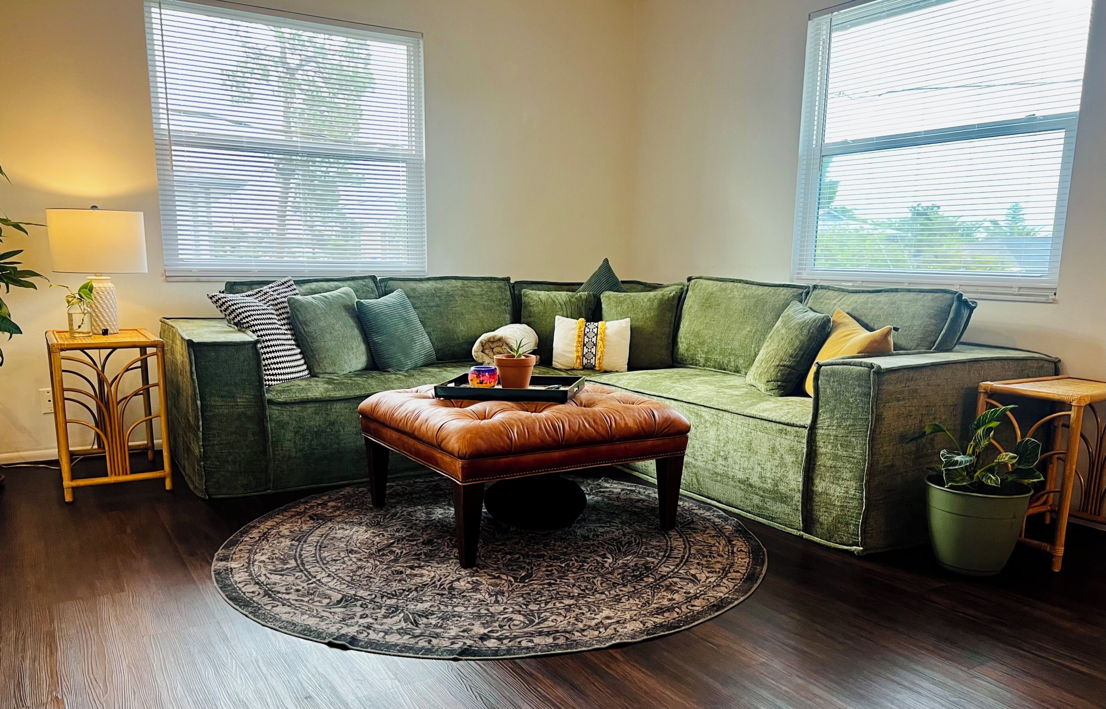 A living room with a green sofa, a brown leather ottoman, a patterned rug, and a small side table with a lamp.