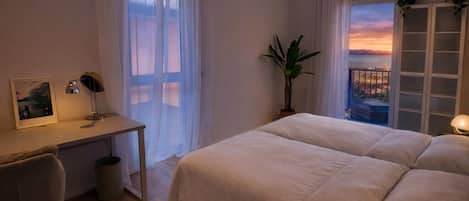 Ein Schlafzimmer mit einem großen Bett, einem Schreibtisch, einem Stuhl und Blick auf den Sonnenuntergang durch eine Balkontür.