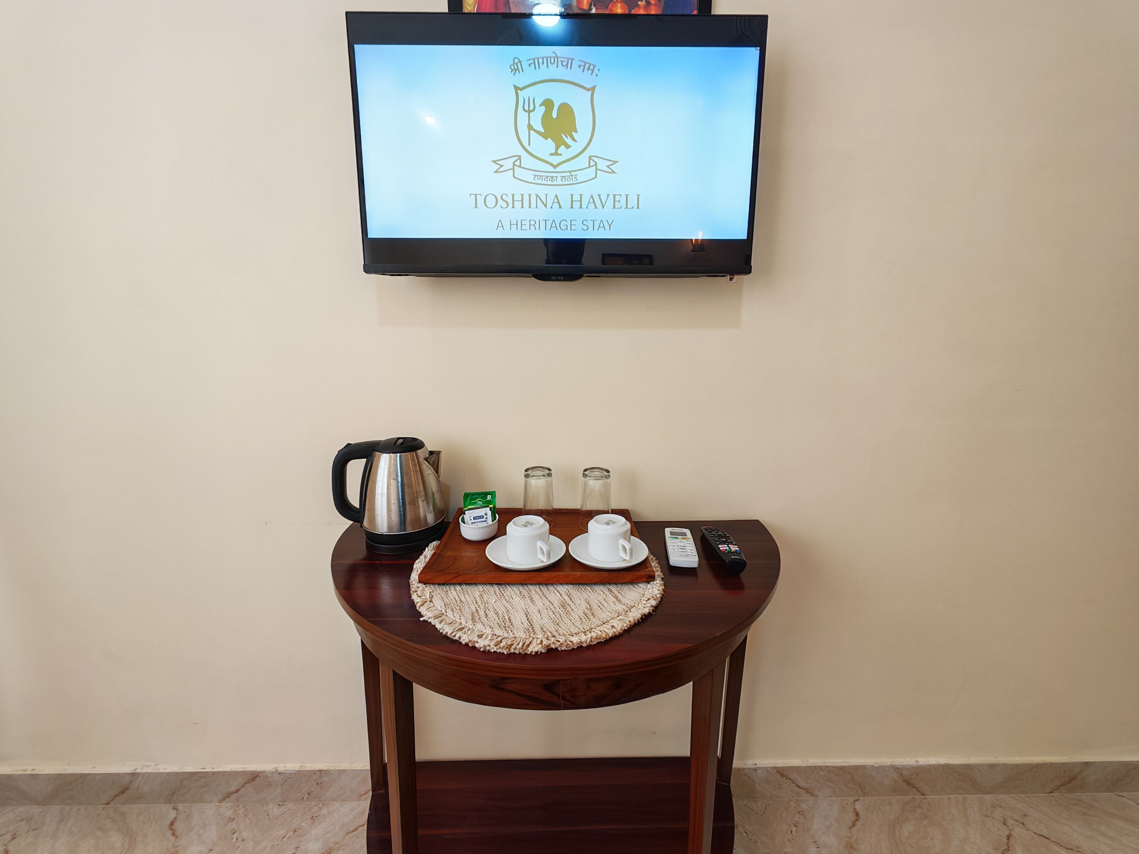 Ein an der Wand befestigter Fernseher zeigt ein Logo und einen Namen. Ein Holztisch mit Wasserkocher, Tassen, einer Fernbedienung und einem Glasbehälter.