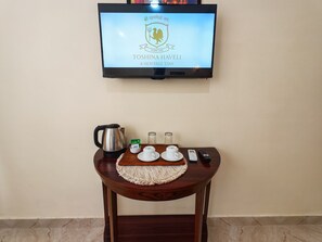 A television mounted on a wall displaying a logo and name, a wooden table with a kettle, cups, a remote, and a glass jar. - Toshina Haveli (Jaipur)