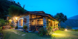 Een stenen huis met een houten veranda, omringd door een verzorgde tuin en een berg op de achtergrond.