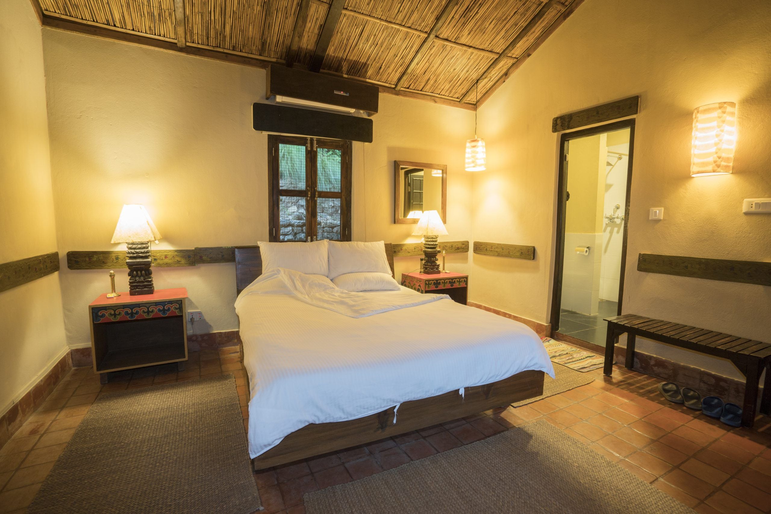 Habitación de hotel con cama, dos mesitas de noche con lámparas, un banco, una ventana y una puerta.