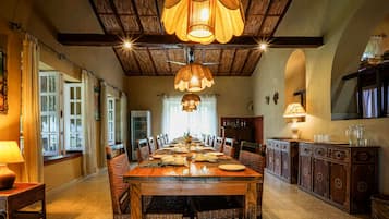 A dining area with a long wooden table set for a meal, flanked by wicker chairs, under a wooden ceiling with exposed beams and hanging lamps.