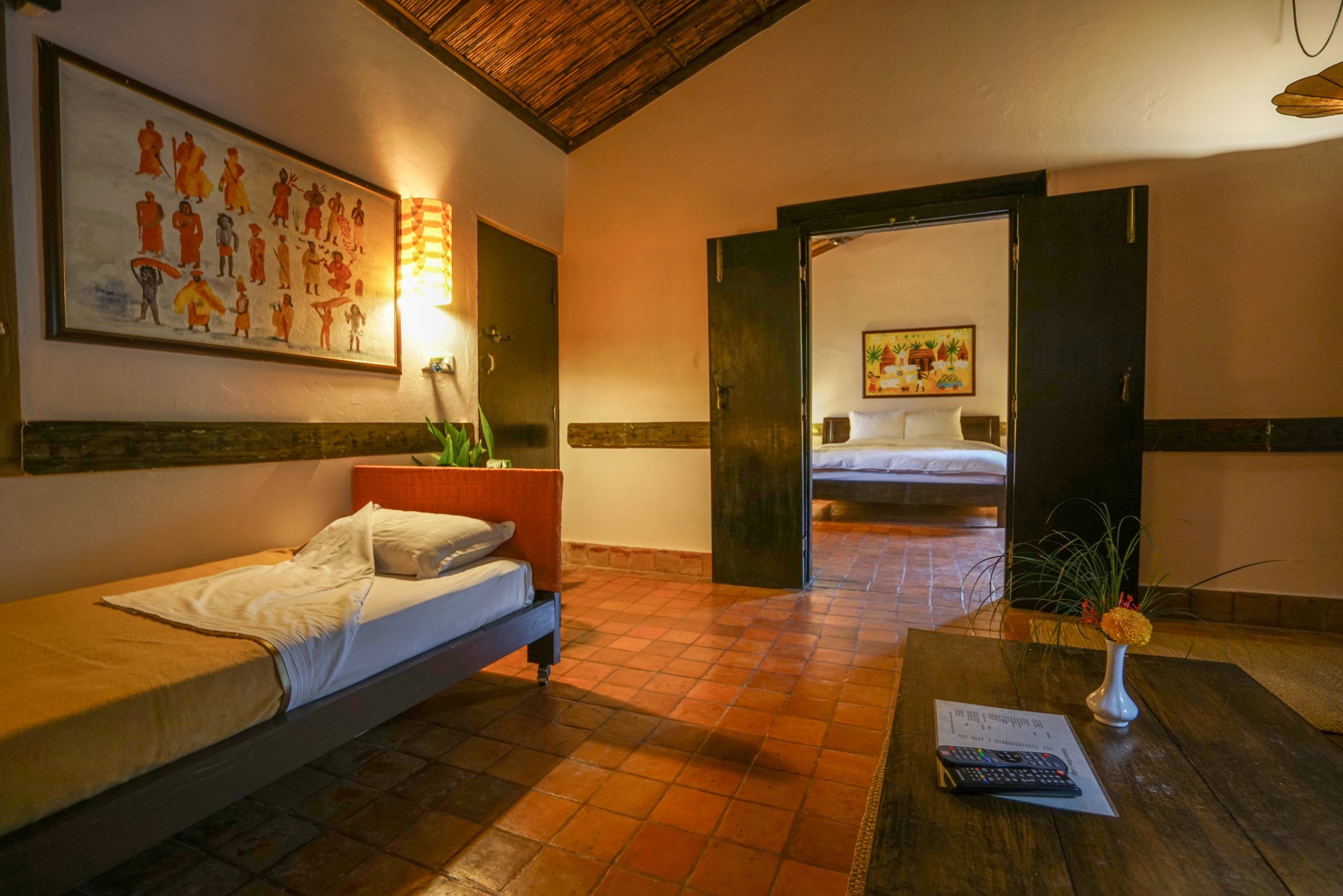 A hotel room with a bed, a desk with a remote control, a plant, and a painting on the wall.