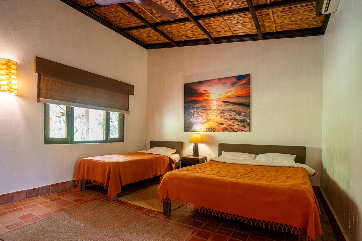 Een hotelkamer met twee bedden, een groot schilderij van een zonsondergang, een raam met uitzicht en een plafondventilator.