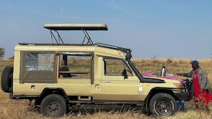 Safariauto, jossa on kattoteline, vararengas sekä pöytä, jonka päällä on vaaleanpunainen liina ja termospullo.