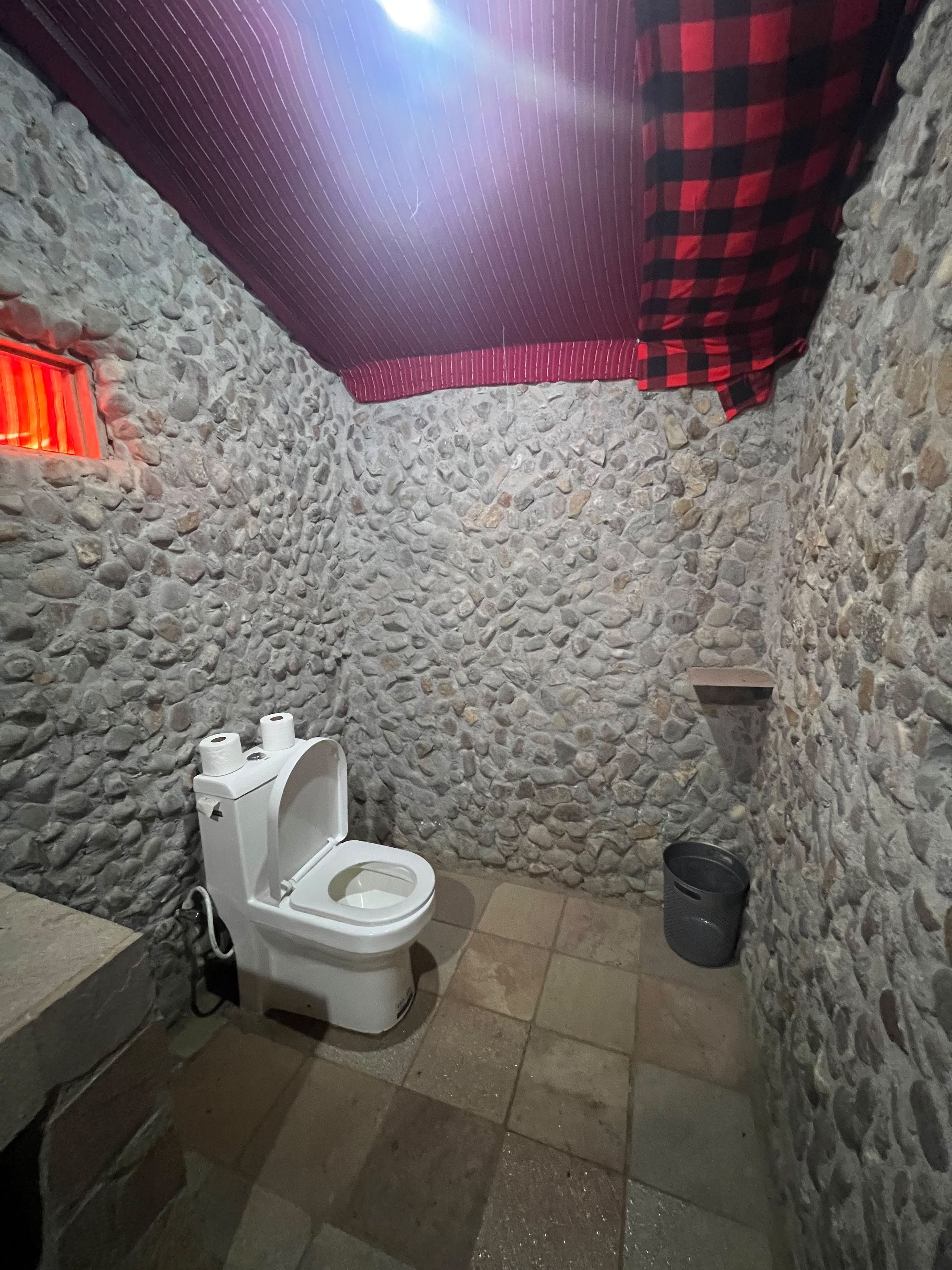 Een betegelde badkamer met een wit toilet, een rood-zwart geruit gordijn en een stenen vloer.
