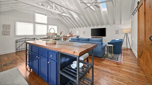 Ein großzügiges Loft mit hoher Decke, einer zentralen Kochinsel mit blauen Schränken, einem blauen Sofa und einem an der Wand montierten Fernseher.