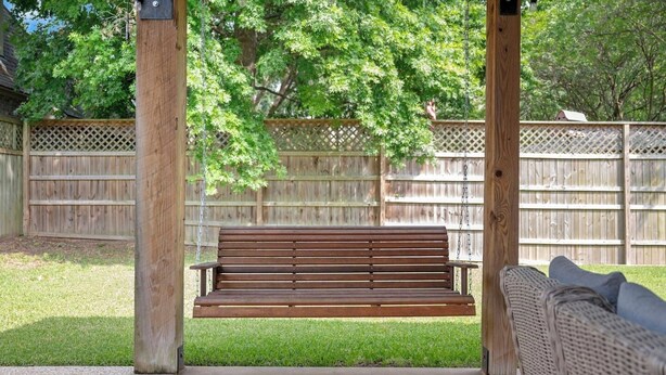 Eine Holzschaukelbank unter einer Holzpergola in einem Garten mit einem Holzzaun im Hintergrund.