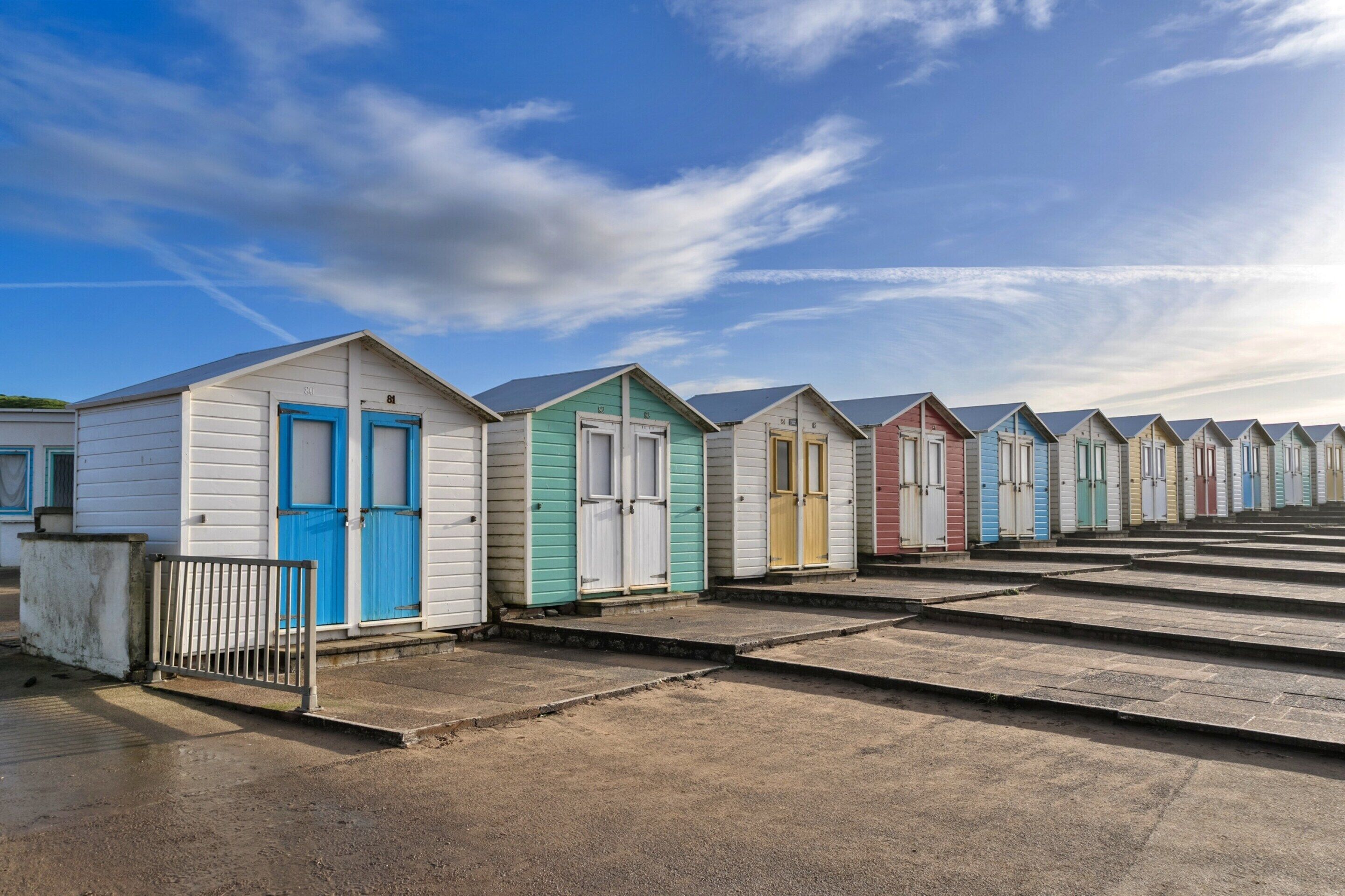 A row of beach huts with numbered doors, arranged in a line along a sandy beach.