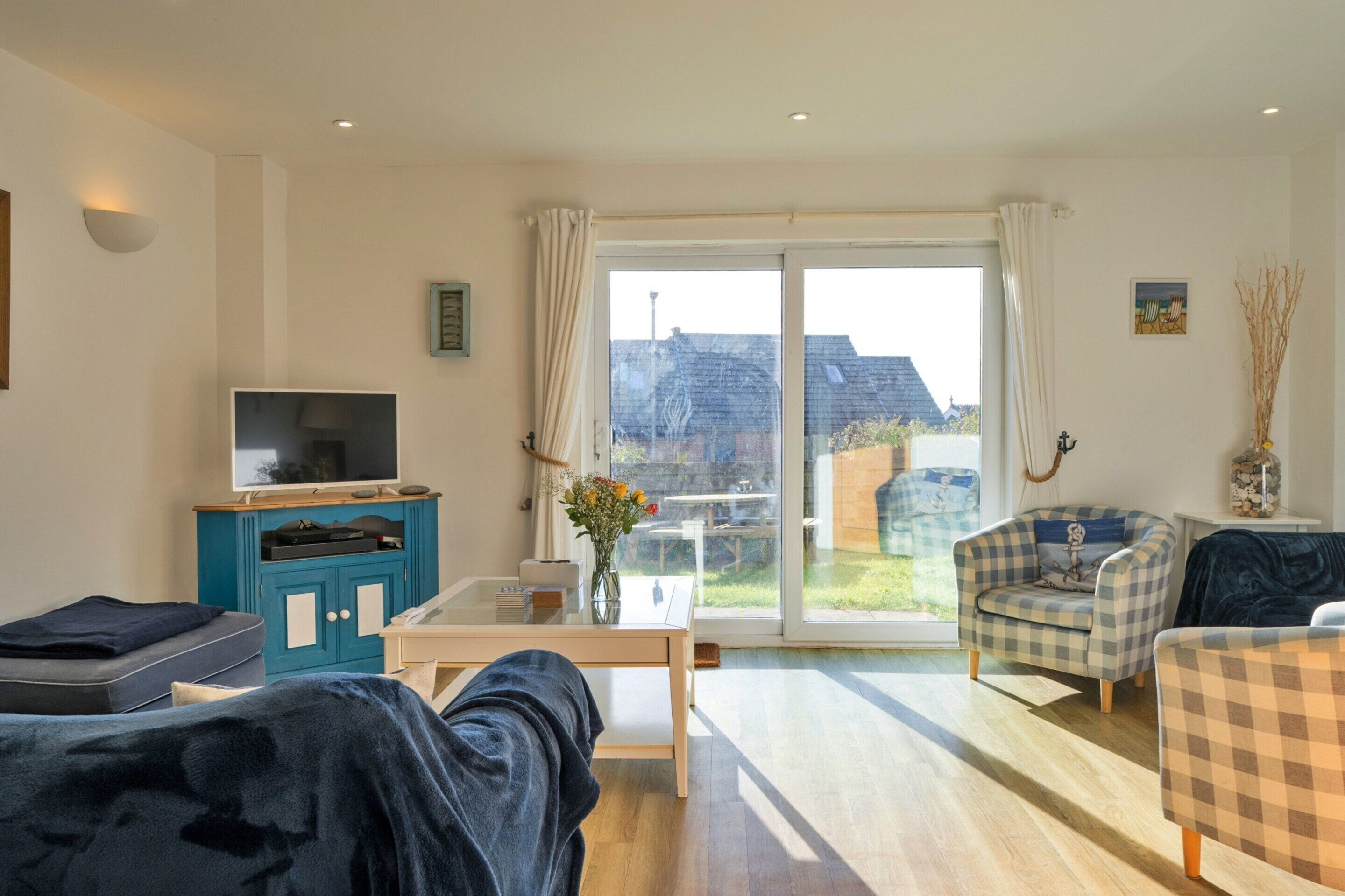 Una sala de estar con televisor, una mesa de centro y dos sillones. Hay un ventanal que da a un balcón con vistas a edificios.