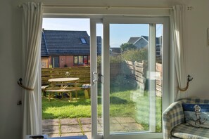 A room with a sliding glass door leading to a patio with a wooden table and chairs, a blue and white checkered sofa, and a view of a house with a red brick wall and a tiled roof. - Bude Sea View Apartment for 4 (Bude)