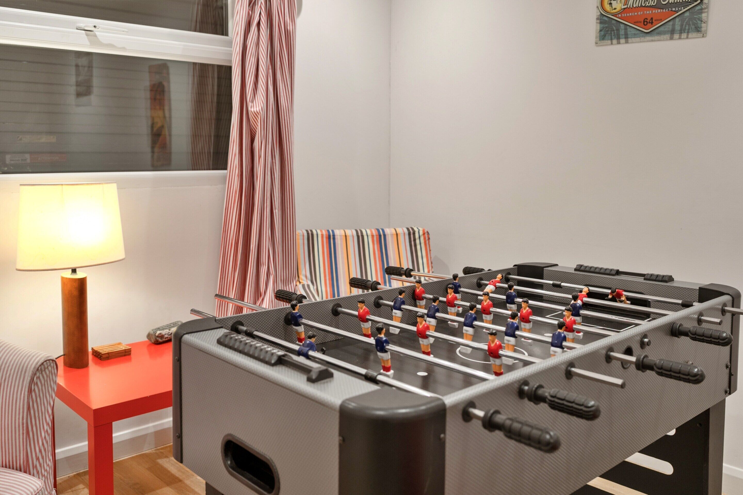 Une table de baby-foot dans une pièce avec une fenêtre, une lampe et des rideaux à rayures.