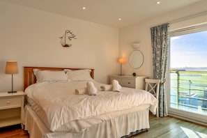 A bedroom with a large bed, a bedside table with a lamp, a small table with a mirror, and a view of the outdoors through a window. - Bude Sea View Apartment for 4 (Bude)