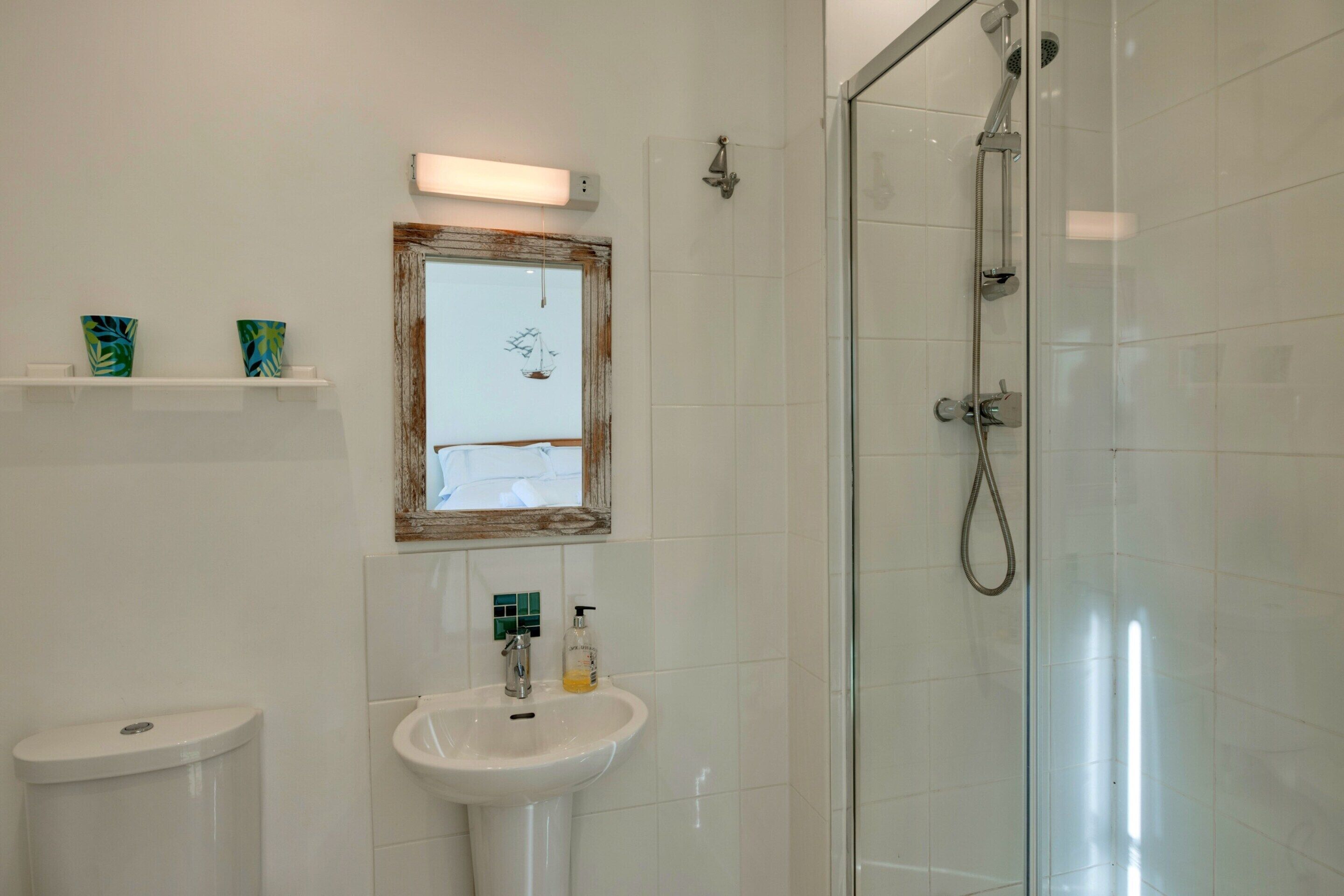 Un baño con cabina de ducha de vidrio, un lavabo blanco y un inodoro. Hay una repisa con objetos decorativos y un cuadro enmarcado sobre el lavabo.