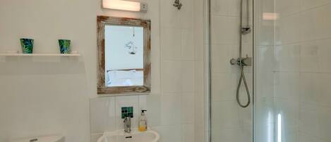 Un baño con cabina de ducha de vidrio, un lavabo blanco y un inodoro. Hay una repisa con objetos decorativos y un cuadro enmarcado sobre el lavabo.