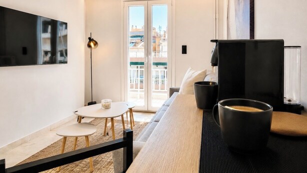 Een moderne woonkamer met een bank, een salontafel, een aan de muur bevestigde televisie en een balkon met uitzicht op gebouwen.