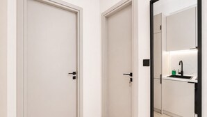 Een gang met twee gesloten deuren en een spiegel die een badkamer weerspiegelt, met een wastafel en een groene zeepdispenser.