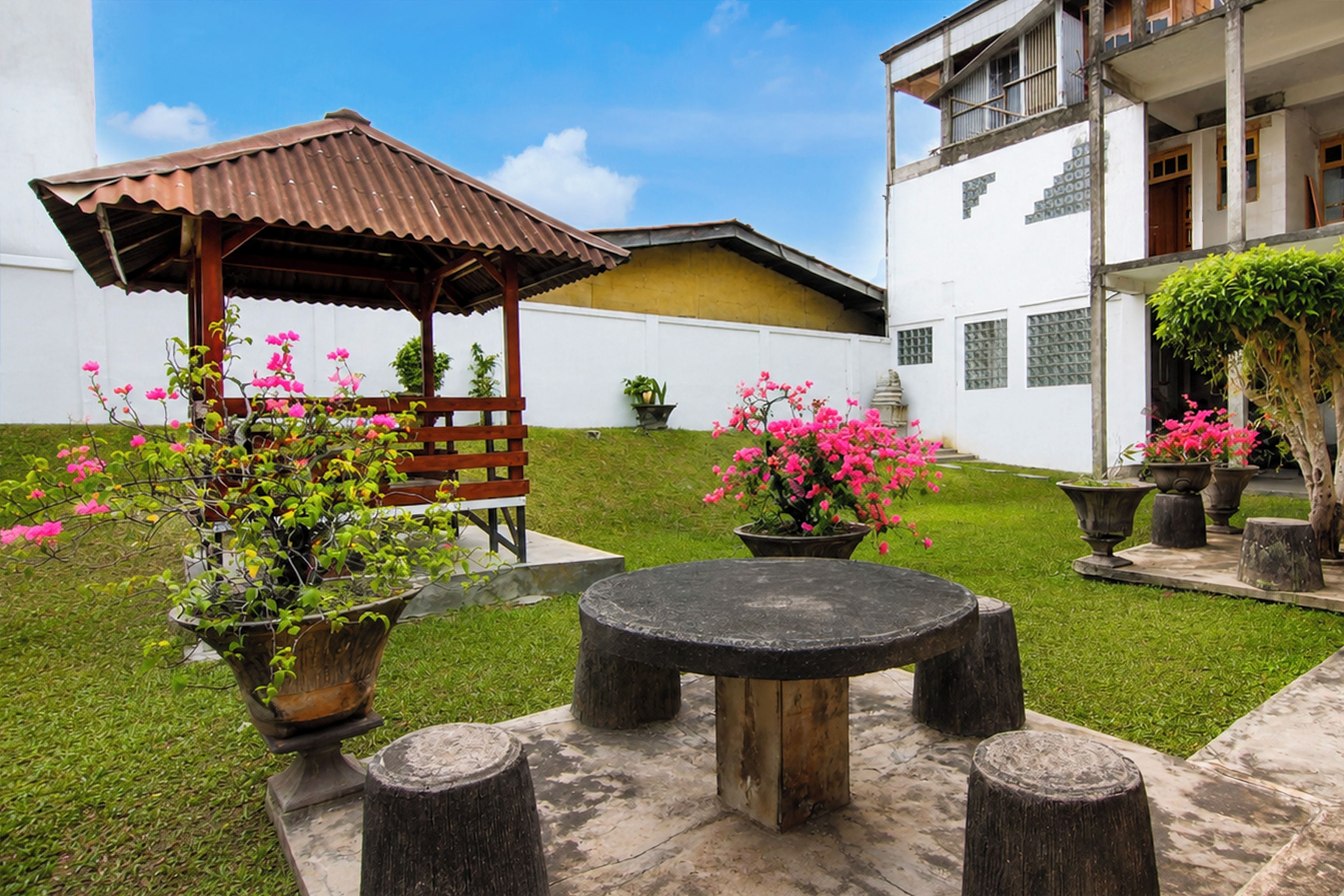 A courtyard with a wooden gazebo, a round table, and potted plants with pink flowers.