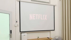 Een kamer met een wandgemonteerde projectiescherm waarop ‘NETFLIX’ in roze letters staat, een klein bureau met een wit apparaat, een staande ventilator en twee houten stoelen.
