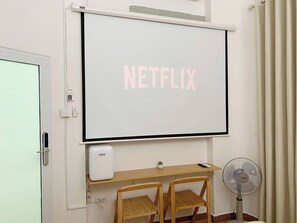 Ein Raum mit einer fest installierten Projektionsleinwand, die 'NETFLIX' in Pink anzeigt, einem kleinen Schreibtisch mit einem weißen Gerät, einem Standventilator und zwei Holzstühlen.