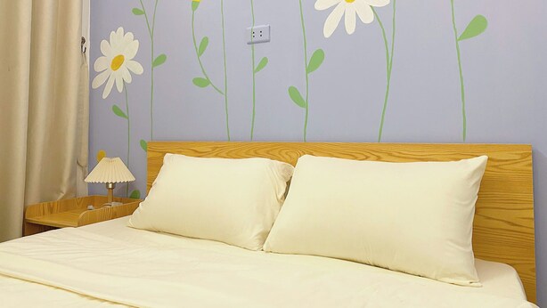 Een slaapkamer met een bed, kussens, een nachtkastje, een lamp en een bloemendecoratie op de muur.