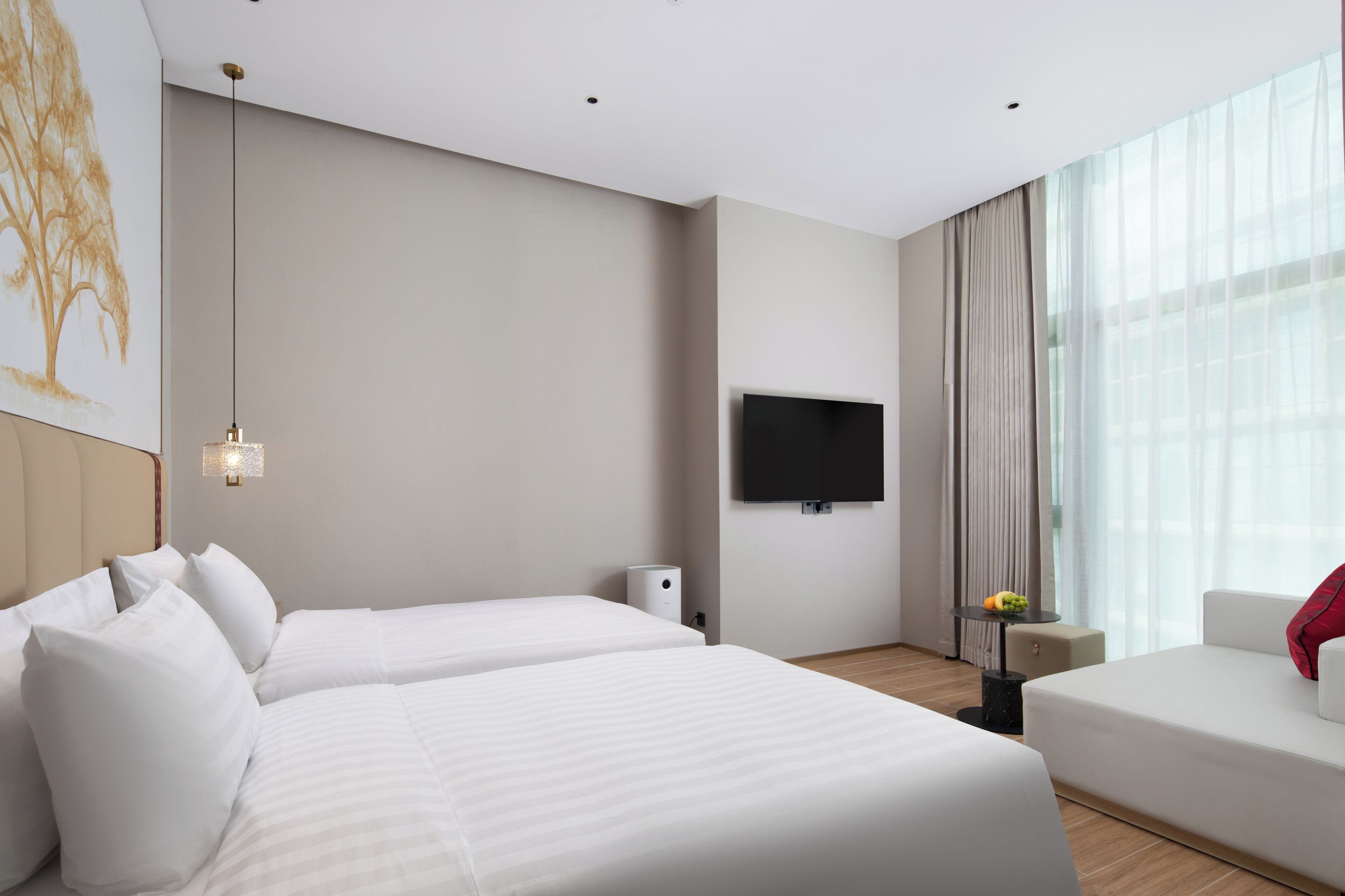 A hotel room with two beds, a flat-screen TV, a small table with a fruit bowl, and a large window with curtains.