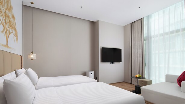 Habitación de hotel con dos camas, un televisor de pantalla plana, una mesita con frutero y un ventanal grande con cortinas.