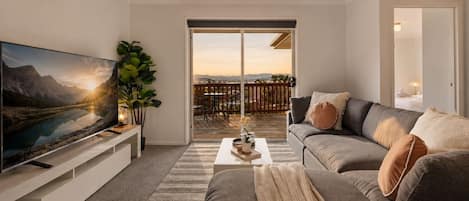 A modern living room with a flat-screen TV, a grey sofa, a coffee table, and a large window overlooking a deck.