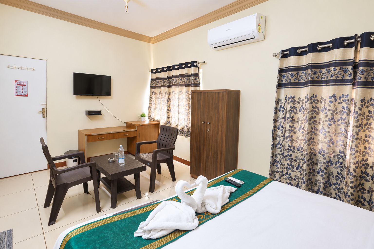 Habitación de hotel con cama, escritorio, dos sillas, televisor, aire acondicionado y cortinas.