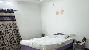 A bedroom with a bed, a nightstand, a wall-mounted outlet, and a curtain. - Hotel The One (Rajkot)