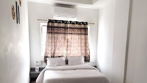 A bedroom with a bed, a nightstand, a window with curtains, and an air conditioning unit. - Hotel The One (Rajkot)