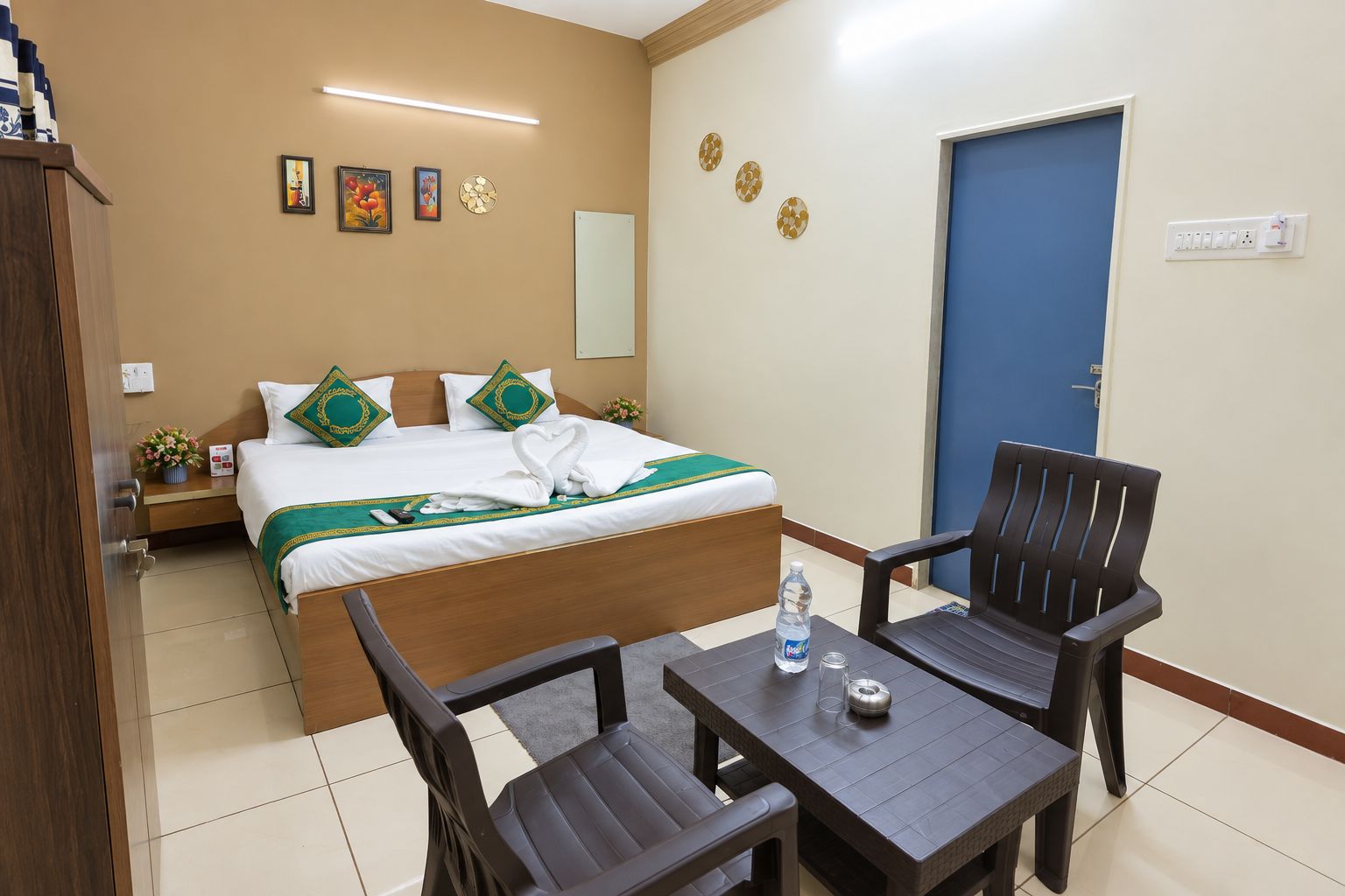 Quarto de hotel com cama, duas cadeiras, mesa, mesinha de cabeceira, relógio de parede, espelho, porta e parede decorada com pratos.