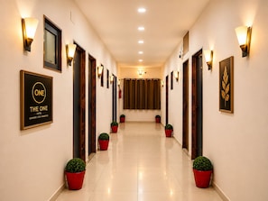 A hotel corridor with framed artwork, wall-mounted lights, and potted plants. - Hotel The One (Rajkot)