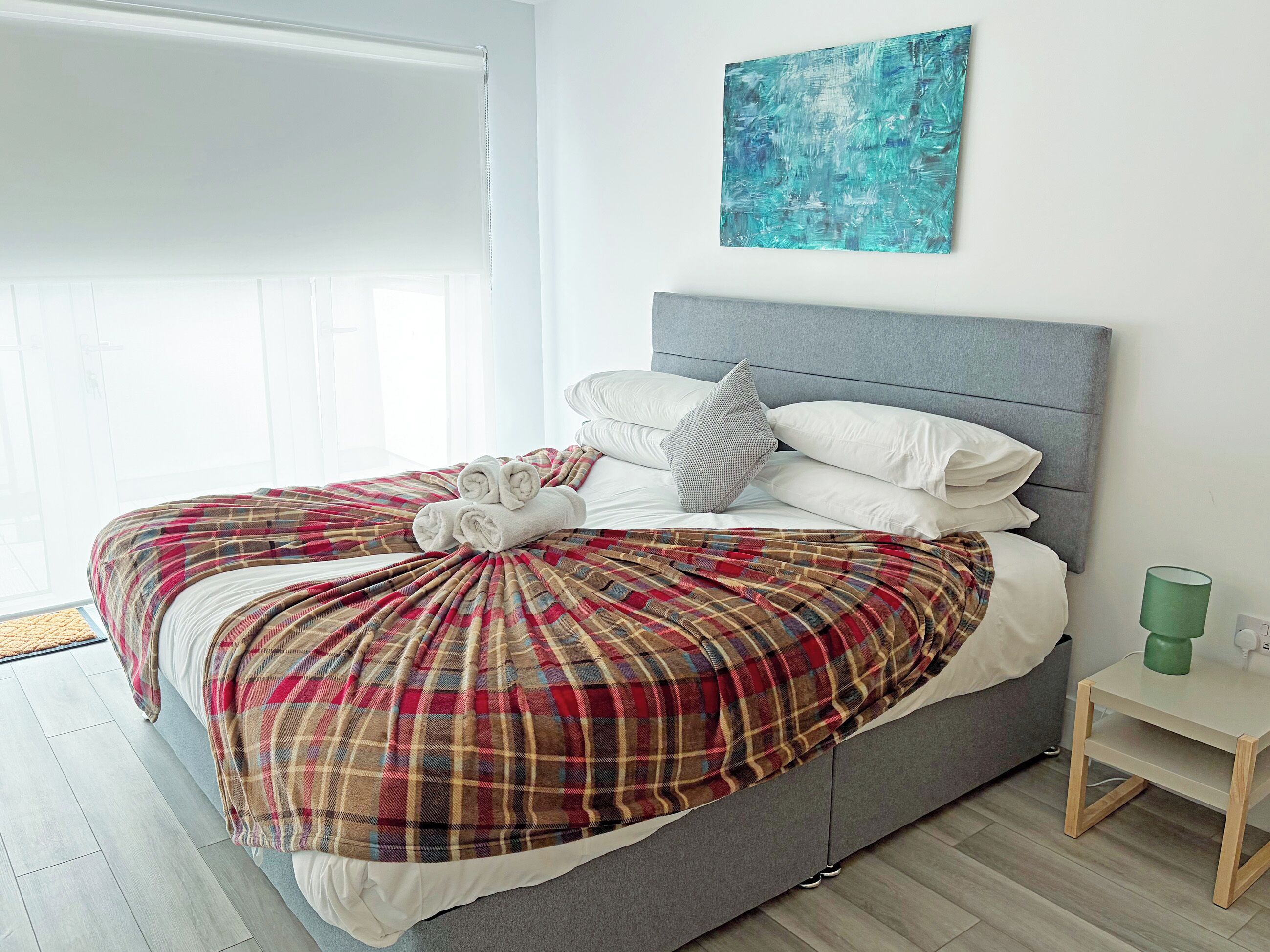 A neatly made bed with a plaid bedspread, a grey headboard, and a green cup on a bedside table.