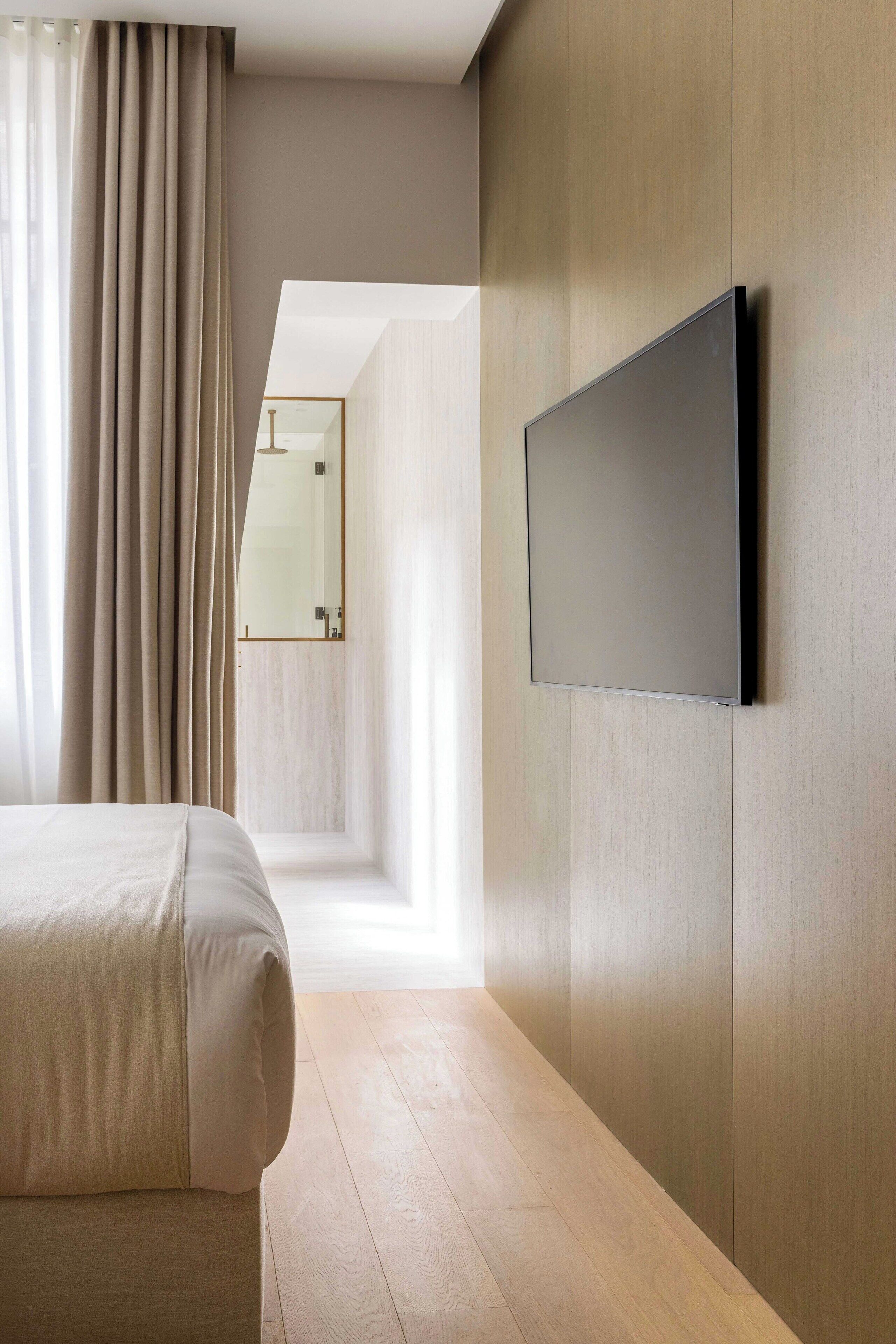 A modern hotel room with a bed, a flat-screen TV mounted on the wall, and a view of a hallway.