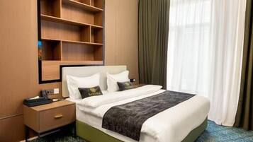 Quarto de hotel com cama, mesas de cabeceira, estante embutida e uma janela grande com cortinas.