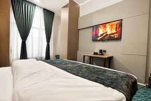 Une chambre d’hôtel avec un lit, un téléviseur à écran plat fixé au mur, une petite table avec une bouilloire et des tasses, et une fenêtre avec des rideaux.