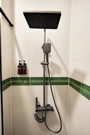 Una ducha con cabezal de ducha de mano y otro fijo en el techo, instalada en una pared con azulejos verdes y blancos.