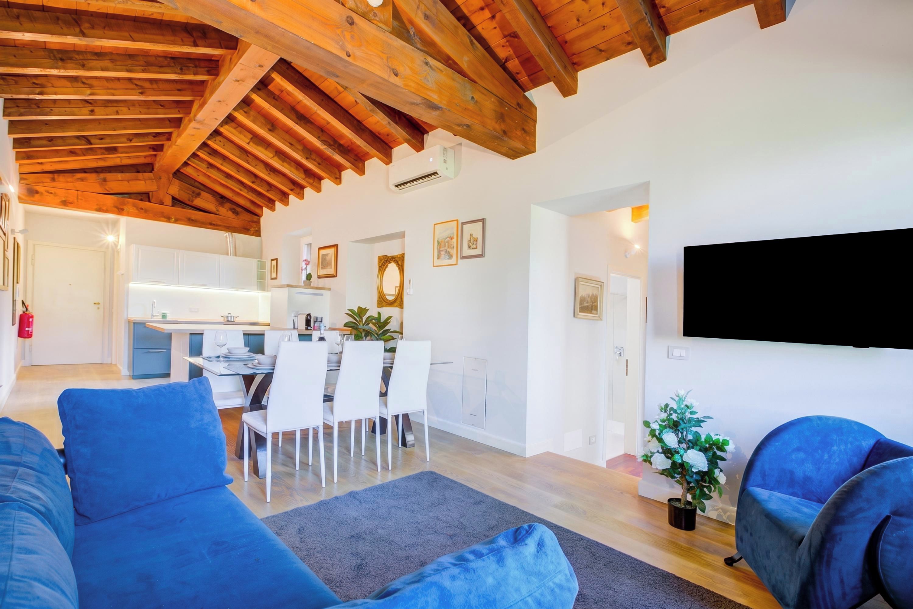 A modern living room with a blue sofa, a dining area with white chairs, and a kitchen with white cabinets.