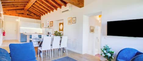 A modern living room with a blue sofa, a dining area with white chairs, and a kitchen with white cabinets.